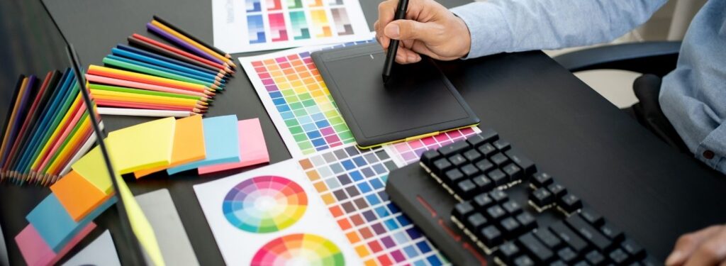 Designer's hands at a keyboard next to printed color swatches and a color wheel