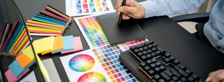 Designer's hands at a keyboard next to printed color swatches and a color wheel